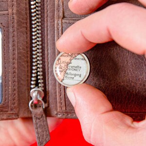 May include: A close-up of a small, round, white map pin with a red outline. The pin shows a map of the Australian coast with the cities of Sydney, Wollongong, Kiama, Parramatta, Lithgow, and Newcastle labelled. The pin is being held by a finger in front of a brown leather wallet.
