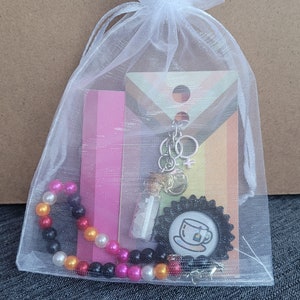 Peut inclure: Un sac en organza blanc rempli d'une variété d'articles, dont une carte rayée rose et blanche, un bracelet en perles noir et blanc, une petite bouteille en verre avec un bouchon en liège et un bouton noir avec un motif de tasse à thé blanche.