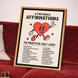 May include: A framed print with the text "A Few Daily Affirmations" and "To Practice Self Love." The artwork features a cartoon heart with arms and legs. The print is displayed on a red velvet couch.
