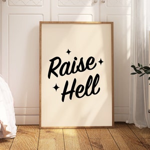 Könnte beinhalten: Ein gerahmter Druck mit weißem Hintergrund und schwarzem Text, der "Raise Hell" mit Sternen auf beiden Seiten des Textes lautet. Der Druck hängt an einer weißen Wand in einem Raum mit einem Bett und einer Vase mit rosa Blumen.