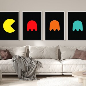 K&ouml;nnte beinhalten: Vier gerahmte Kunstdrucke mit Retro-Videospielcharakteren. Die Drucke zeigen einen gelben Pac-Man und drei farbige Geister auf schwarzem Hintergrund. Das Kunstwerk wird &uuml;ber einem wei&szlig;en Sofa ausgestellt.