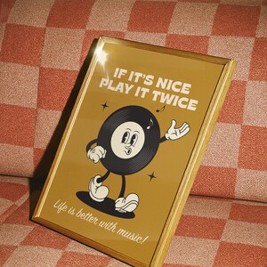Puede incluir: Un estampado enmarcado con un borde dorado que presenta un personaje de disco de vinilo de dibujos animados. El personaje tiene una cara sonriente y gesticula con una mano. El texto dice "IF IT'S NICE PLAY IT TWICE" y "Life is better with music!"