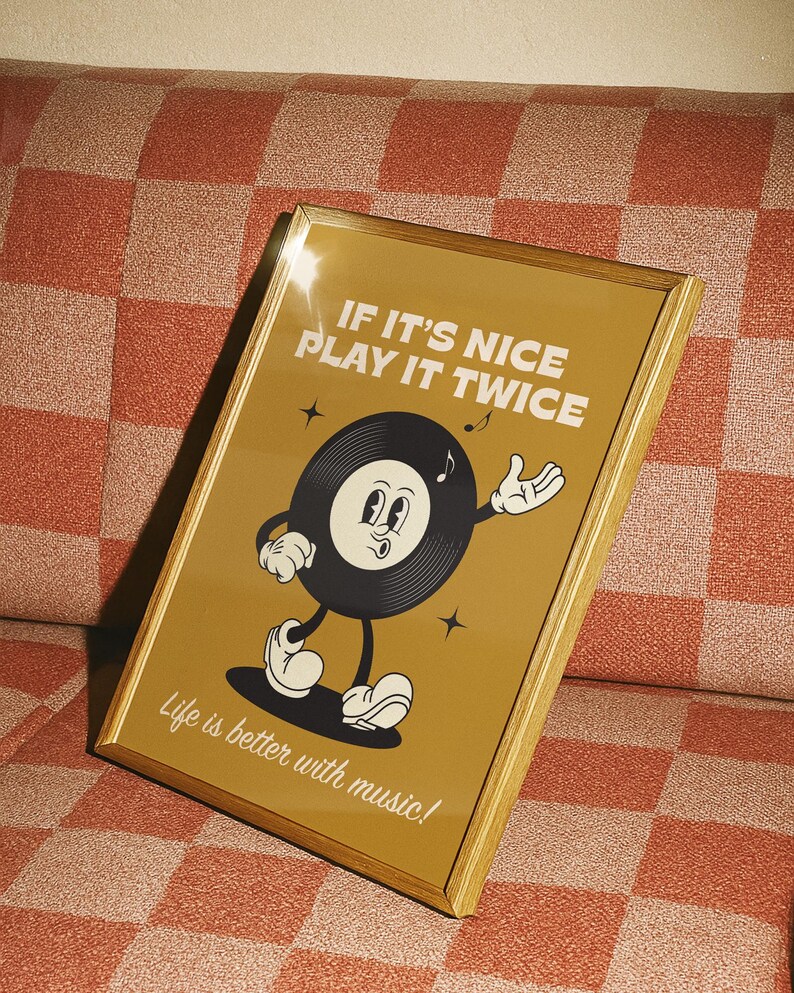 Puede incluir: Un estampado enmarcado con un borde dorado que presenta un personaje de disco de vinilo de dibujos animados. El personaje tiene una cara sonriente y gesticula con una mano. El texto dice "IF IT'S NICE PLAY IT TWICE" y "Life is better with music!"