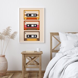 May include: A framed print of three vintage cassette tapes with a beige background. The tapes are stacked vertically and have a retro design with orange, brown, and pink accents. The print is hanging on a white wall above a wooden nightstand with a book and a white mug.