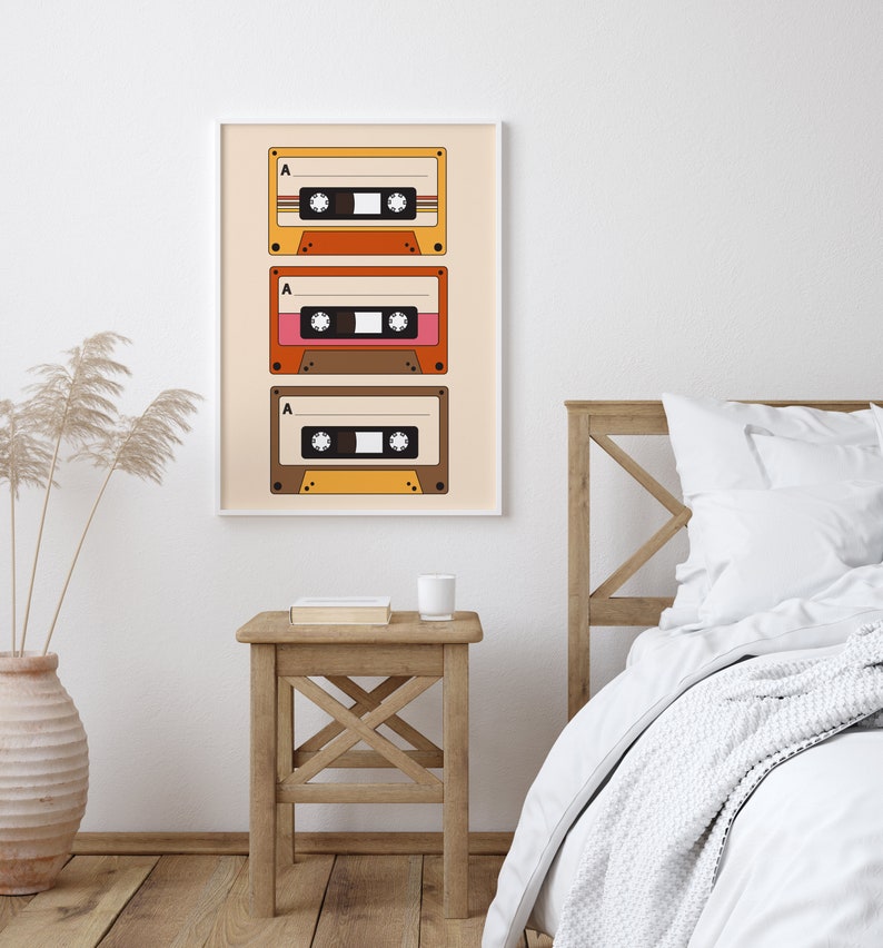 May include: A framed print of three vintage cassette tapes with a beige background. The tapes are stacked vertically and have a retro design with orange, brown, and pink accents. The print is hanging on a white wall above a wooden nightstand with a book and a white mug.