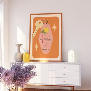 May include: A framed print of a pink motel key with a palm tree and the text "Palm Canyon Motel & Resort 1989 Palm Springs, CA". The print has a light orange background with white stars.