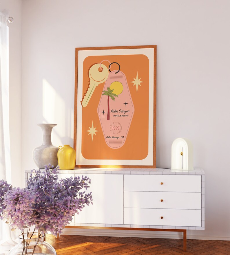 May include: A framed print of a pink motel key with a palm tree and the text "Palm Canyon Motel & Resort 1989 Palm Springs, CA". The print has a light orange background with white stars.