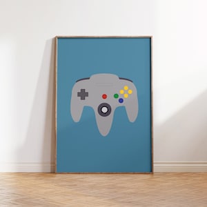 May include: A framed poster with a blue background and a gray Nintendo 64 controller in the center. The controller has a red button, green button, and yellow buttons.