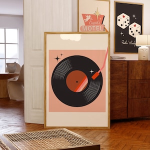 May include: A pink and black retro-style print of a vinyl record with a red needle. The print is framed in a light wood frame and is hanging on a white wall. The print is surrounded by other framed prints, including one that says "Cupid's Motel" and another that says "Feelin' Lucky".
