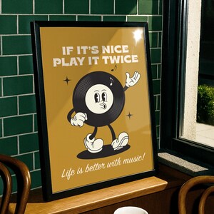 Puede incluir: Un p&oacute;ster enmarcado con un personaje de disco de vinilo de dibujos animados retro sobre un fondo amarillo mostaza. El texto dice "IF IT'S NICE PLAY IT TWICE" y "Life is better with music!". El p&oacute;ster se muestra en una repisa de madera.
