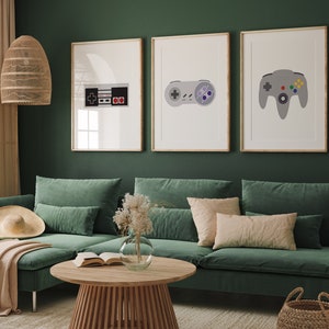 Puede incluir: Tres impresiones enmarcadas de mandos de videojuegos retro y un cartucho de juego. Las impresiones est&aacute;n en una pared verde sobre un sof&aacute; verde con almohadas y una manta.