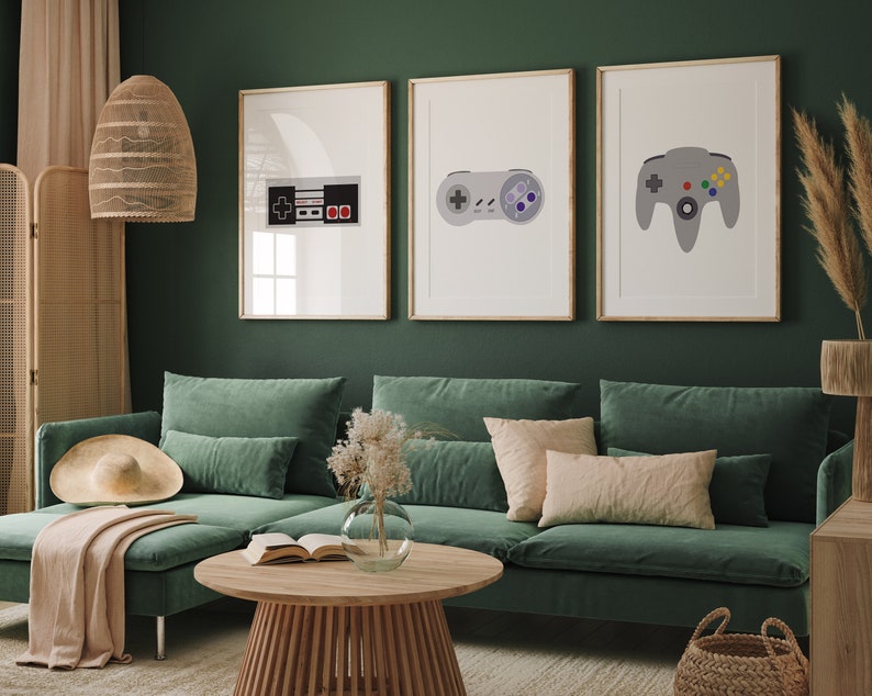 Puede incluir: Tres impresiones enmarcadas de mandos de videojuegos retro y un cartucho de juego. Las impresiones est&aacute;n en una pared verde sobre un sof&aacute; verde con almohadas y una manta.