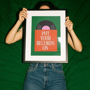 May include: A framed art print featuring a vinyl record design. The artwork has a green background with an orange rectangle at the bottom. The text "PUT YOUR RECORDS ON" is printed in white. The record is black with a pink center.