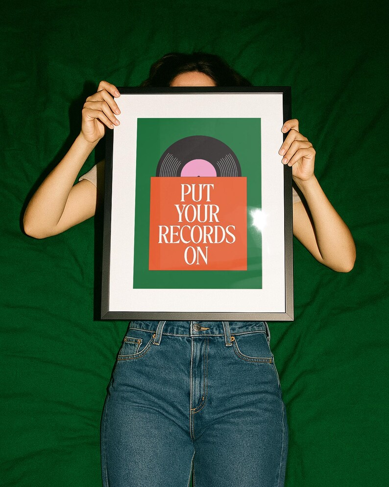 May include: A framed art print featuring a vinyl record design. The artwork has a green background with an orange rectangle at the bottom. The text "PUT YOUR RECORDS ON" is printed in white. The record is black with a pink center.