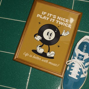 Puede incluir: Un estampado enmarcado con un personaje de disco de vinilo de dibujos animados sobre un fondo amarillo mostaza. El texto dice "IF IT'S NICE PLAY IT TWICE" y "Life is better with music!". Una zapatilla azul y blanca est&aacute; al lado.