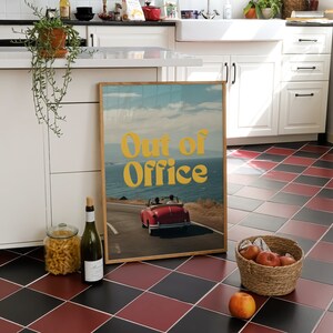 May include: A framed print with a retro aesthetic featuring a red convertible car driving on a winding road with a blue ocean and sky in the background. The text "Out of Office" is in yellow letters on the print.