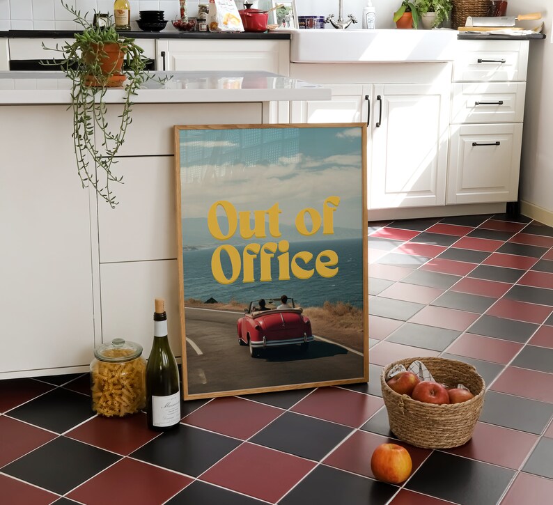 May include: A framed print with a retro aesthetic featuring a red convertible car driving on a winding road with a blue ocean and sky in the background. The text "Out of Office" is in yellow letters on the print.