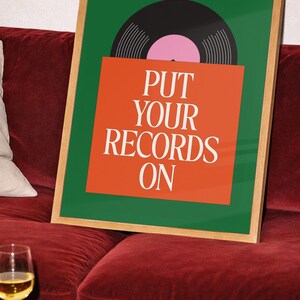 May include: A framed art print with a green background and a black vinyl record graphic. The print features the text "PUT YOUR RECORDS ON" in white on an orange background. The frame is a light wood color.