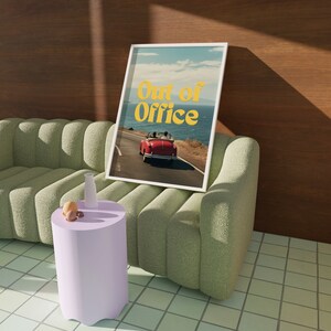 May include: A white framed print with the text "Out of Office" in yellow letters. The print features a red convertible car driving on a coastal road with a blue ocean in the background.