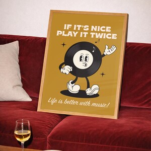Puede incluir: Un p&oacute;ster enmarcado con un dise&ntilde;o de disco de vinilo de dibujos animados vintage. El p&oacute;ster dice "IF IT'S NICE PLAY IT TWICE" y "Life is better with music!". El personaje del disco tiene una cara sonriente y est&aacute; gesticulando. El p&oacute;ster est&aacute; sobre un sof&aacute; de terciopelo rojo.