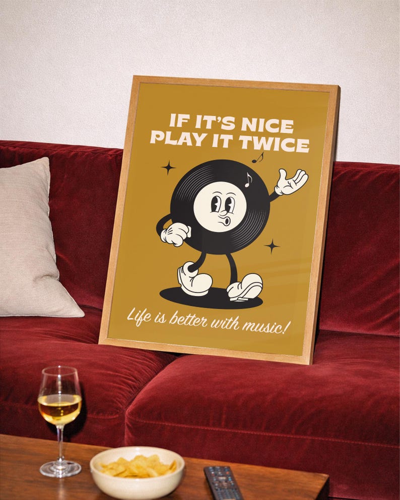 Puede incluir: Un p&oacute;ster enmarcado con un dise&ntilde;o de disco de vinilo de dibujos animados vintage. El p&oacute;ster dice "IF IT'S NICE PLAY IT TWICE" y "Life is better with music!". El personaje del disco tiene una cara sonriente y est&aacute; gesticulando. El p&oacute;ster est&aacute; sobre un sof&aacute; de terciopelo rojo.