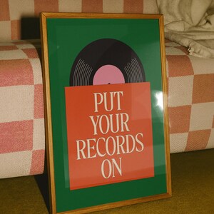 May include: A framed art print featuring a vinyl record design. The artwork has a green background with a black record and pink center. The text "PUT YOUR RECORDS ON" is displayed on a red rectangle. The frame is made of wood.