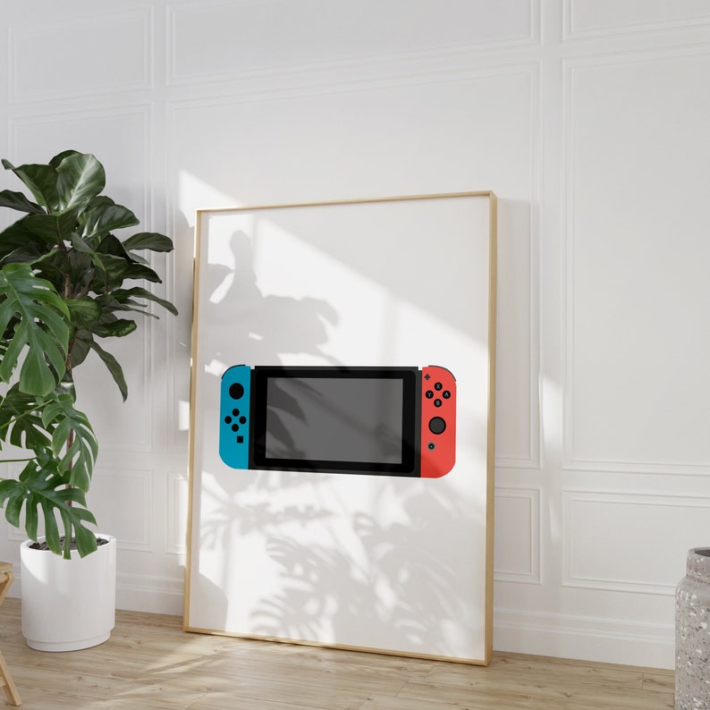 Puede incluir: Una imagen enmarcada en blanco con una sombra de una planta sobre ella. La imagen muestra una consola Nintendo Switch azul y roja con una pantalla negra.