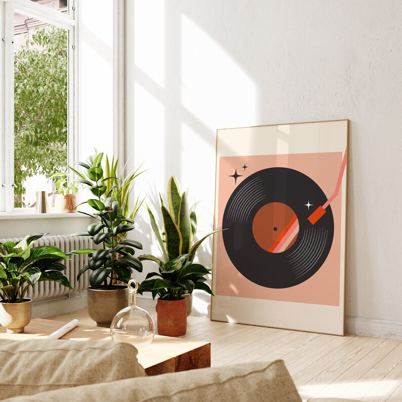 May include: A framed print of a black vinyl record with a pink and orange needle on a pink background. The print is hanging on a white wall in a room with plants and a window.