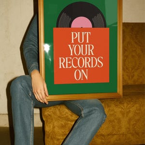 May include: A framed art print featuring a vinyl record design. The artwork has a green background with a black and pink record and an orange square with the text "PUT YOUR RECORDS ON" in white. The frame is gold.