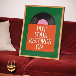 May include: A framed art print with a green background and a vinyl record illustration. The print features the text "PUT YOUR RECORDS ON" in white on an orange background. The artwork is displayed on a red velvet couch, with a glass of wine and snacks on a table.