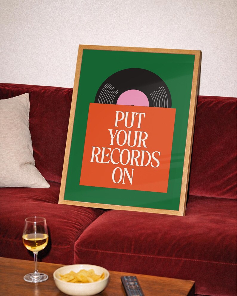 May include: A framed art print with a green background and a vinyl record illustration. The print features the text "PUT YOUR RECORDS ON" in white on an orange background. The artwork is displayed on a red velvet couch, with a glass of wine and snacks on a table.