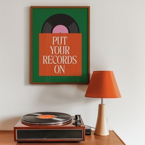 May include: A framed art print with the text "PUT YOUR RECORDS ON" above a record player and lamp. The print has a green background with a black and pink record design. The lamp has an orange shade.