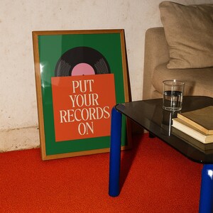 May include: A framed art print with a green background and a vinyl record graphic. The print has the text "PUT YOUR RECORDS ON" in white on an orange background. The frame is brown and the print is on a red carpet.