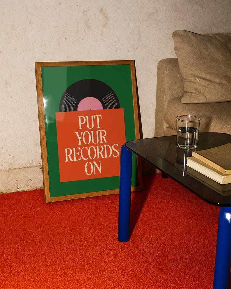 May include: A framed art print with a green background and a vinyl record graphic. The print has the text "PUT YOUR RECORDS ON" in white on an orange background. The frame is brown and the print is on a red carpet.