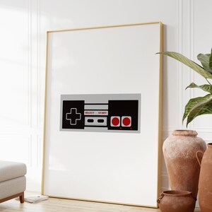 Puede incluir: Una impresi&oacute;n enmarcada de un mando de videojuego retro con botones negros y grises y detalles rojos. El mando tiene las palabras "Select" y "Start" impresas en &eacute;l.