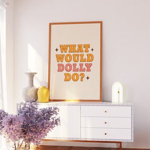 K&ouml;nnte beinhalten: Ein gerahmter Druck mit dem Text "WHAT WOULD DOLLY DO?" in orangefarbenen und rosafarbenen Buchstaben auf wei&szlig;em Hintergrund. Der Druck h&auml;ngt an einer wei&szlig;en Wand &uuml;ber einer wei&szlig;en Kommode mit goldenen Griffen. Auf der Kommode stehen eine Vase mit lila Blumen und eine gelbe Vase.