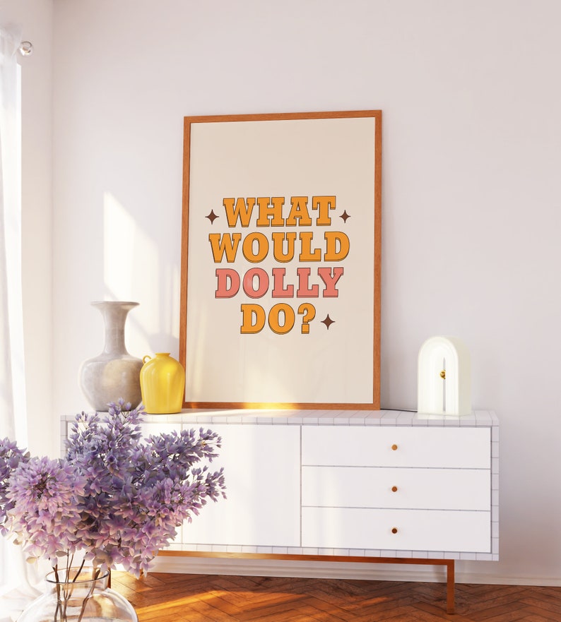K&ouml;nnte beinhalten: Ein gerahmter Druck mit dem Text "WHAT WOULD DOLLY DO?" in orangefarbenen und rosafarbenen Buchstaben auf wei&szlig;em Hintergrund. Der Druck h&auml;ngt an einer wei&szlig;en Wand &uuml;ber einer wei&szlig;en Kommode mit goldenen Griffen. Auf der Kommode stehen eine Vase mit lila Blumen und eine gelbe Vase.
