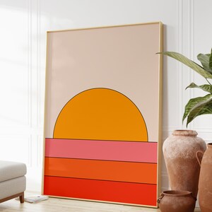 May include: A framed abstract art print with a sunset design. The print features a large orange circle representing the sun, with a pink, orange, and red striped background.