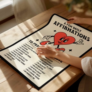 May include: A poster with a black border and a cream background featuring a red heart character with arms and legs. The poster has the text "A Few Daily Affirmations" and a list of positive affirmations. The poster is on a wooden table.