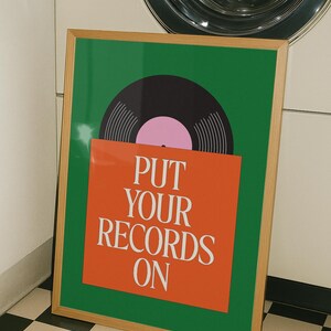 May include: A framed poster with a green background and a vinyl record graphic. The record is black with a pink center. An orange rectangle at the bottom has the text "PUT YOUR RECORDS ON" in white.