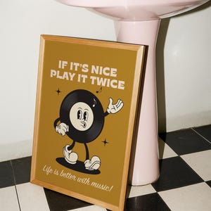 Puede incluir: Un p&oacute;ster enmarcado con un personaje de disco de vinilo de dibujos animados sobre un fondo amarillo mostaza. El texto dice "IF IT'S NICE PLAY IT TWICE" y "Life is better with music!". El marco es de color marr&oacute;n claro.