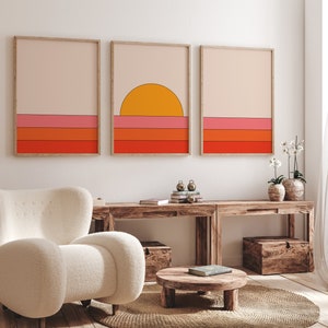 May include: Three framed prints with a minimalist geometric design featuring a sunset over a horizon with orange, pink, and red stripes. The prints are hanging on a white wall above a wooden table with a white armchair and a small wooden table.