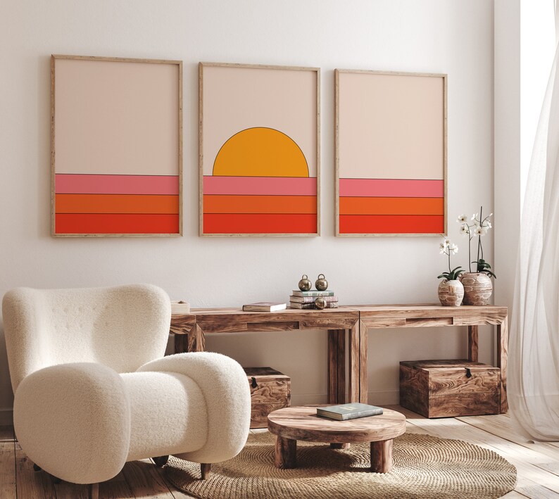 May include: Three framed prints with a minimalist geometric design featuring a sunset over a horizon with orange, pink, and red stripes. The prints are hanging on a white wall above a wooden table with a white armchair and a small wooden table.