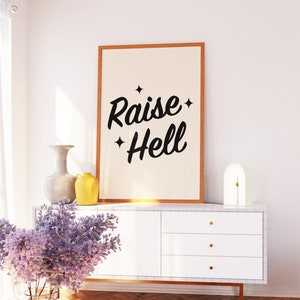 May include: A white dresser with three drawers and gold handles. A framed print with the text "Raise Hell" hangs above the dresser. A vase with purple flowers sits on the floor in front of the dresser. A yellow vase and a gray vase sit on top of the dresser.