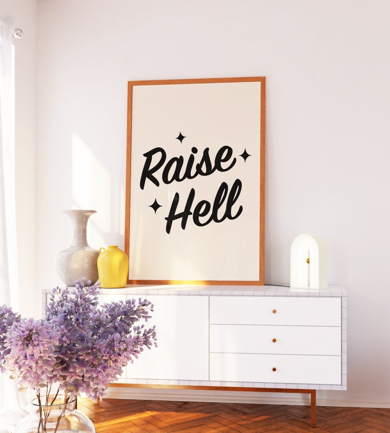 May include: A white dresser with three drawers and gold handles. A framed print with the text "Raise Hell" hangs above the dresser. A vase with purple flowers sits on the floor in front of the dresser. A yellow vase and a gray vase sit on top of the dresser.