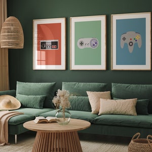 May include: Three framed prints of retro video game controllers on a green wall above a green sectional sofa with pillows. The prints feature a red, green, and blue background with a black and white NES controller, a gray Sega Genesis controller, and a gray Nintendo 64 controller.