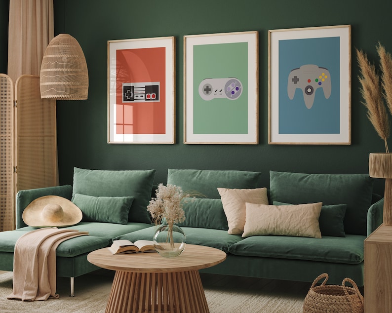 May include: Three framed prints of retro video game controllers on a green wall above a green sectional sofa with pillows. The prints feature a red, green, and blue background with a black and white NES controller, a gray Sega Genesis controller, and a gray Nintendo 64 controller.