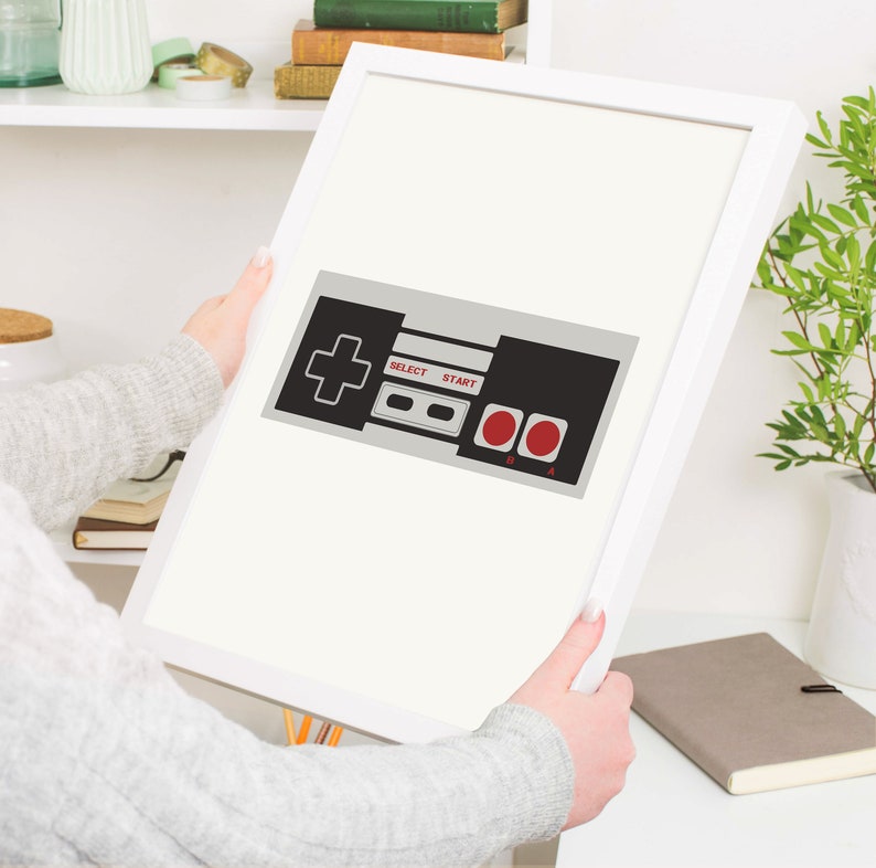 Puede incluir: Una impresi&oacute;n enmarcada de un mando de videojuego vintage con botones negros y grises, un signo m&aacute;s rojo y botones de inicio y selecci&oacute;n rojos. El texto "SELECT" y "START" est&aacute; impreso en los botones.