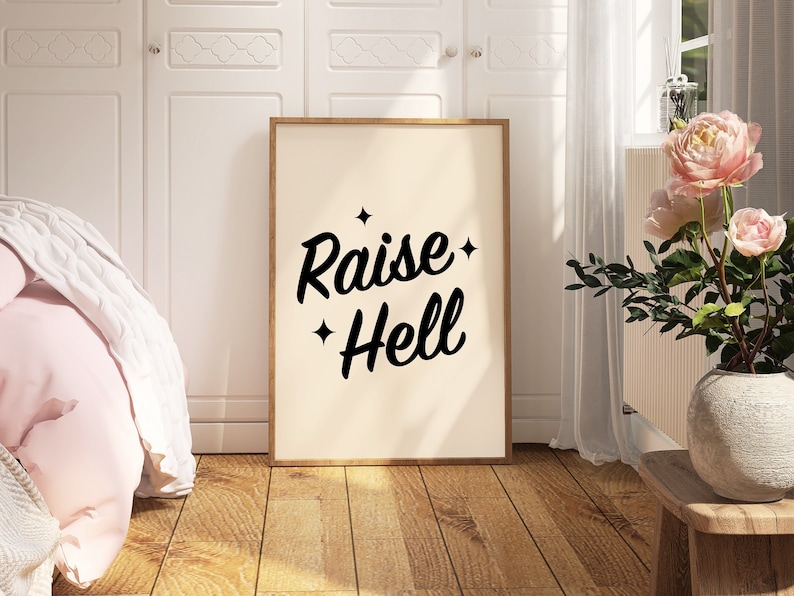 May include: A framed print with a white background and black text that reads "Raise Hell" with stars on either side of the text. The print is hanging on a white wall in a room with a bed and a vase of pink flowers.