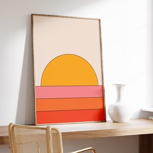 May include: A framed print with a minimalist geometric design of a yellow sun setting over a horizon with pink, orange, and red stripes.